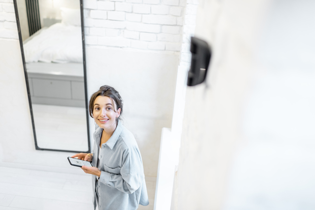 Woman controlling alarm system with a smart phone wireless, standing in the room with motion sensor mounted on the wall